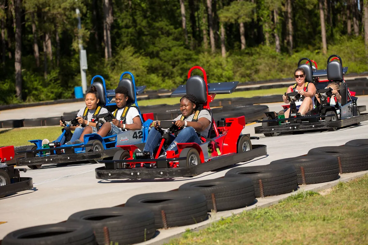 A dynamic scene showing families and friends racing go-karts on a winding outdoor track. The excitement is palpable, with drivers focused and tire barriers marking the course. Spectators and racers create an energetic, playful environment that emphasizes shared adventures and competitive fun.