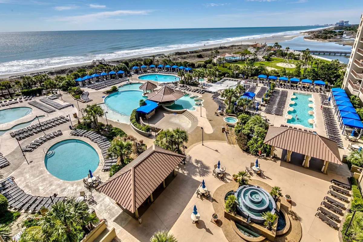 This vibrant exterior image captures sprawling swimming pools bordered by palm trees and ample lounge chairs, all set against a beachfront backdrop. A gazebo is visible, and guests can be seen swimming, sunbathing, and socializing, highlighting a classic resort pool oasis atmosphere.