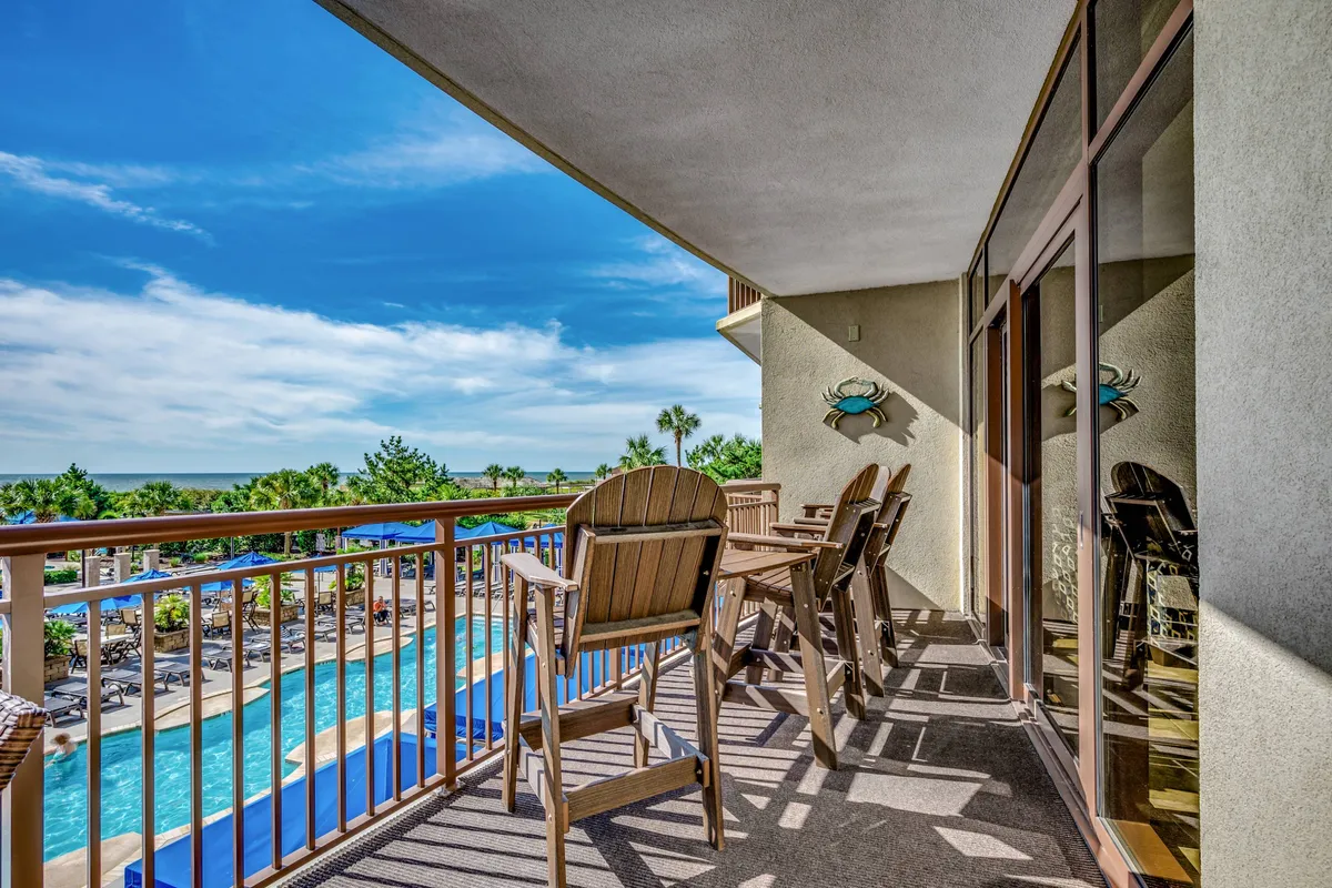 The image showcases a serene condo balcony with inviting seating, overlooking a sparkling pool and lush coastal surroundings. Reflections on the glass and soft lighting evoke a peaceful, sunset mood, hinting at relaxing mornings and tranquil evenings.