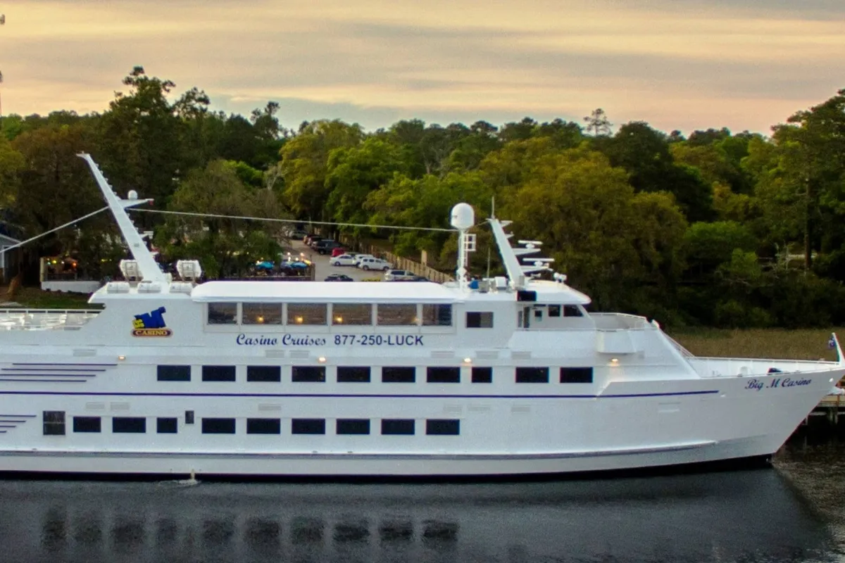 A picturesque cruise ship glides over calm waters amid a backdrop of lush trees and open sky. The ship is aglow, indicating an atmosphere of excitement and luxury, ideal for dining, entertainment, and waterfront exploration.