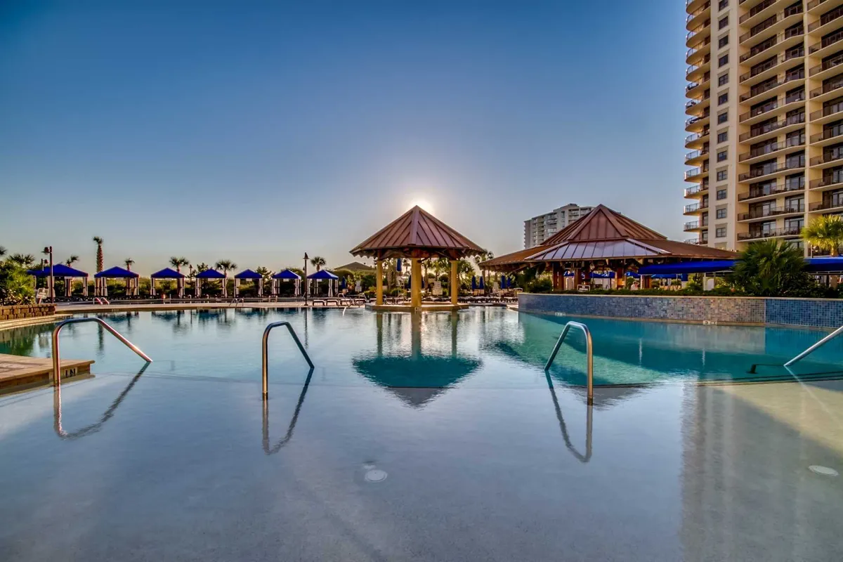 A lush, tropical poolside setting featuring a sparkling swimming pool bordered by palm trees and stylish cabanas, all set against a vibrant sunset sky. Groups of people can be seen socializing and relaxing by the pool, creating an inviting atmosphere for shared outdoor fun.