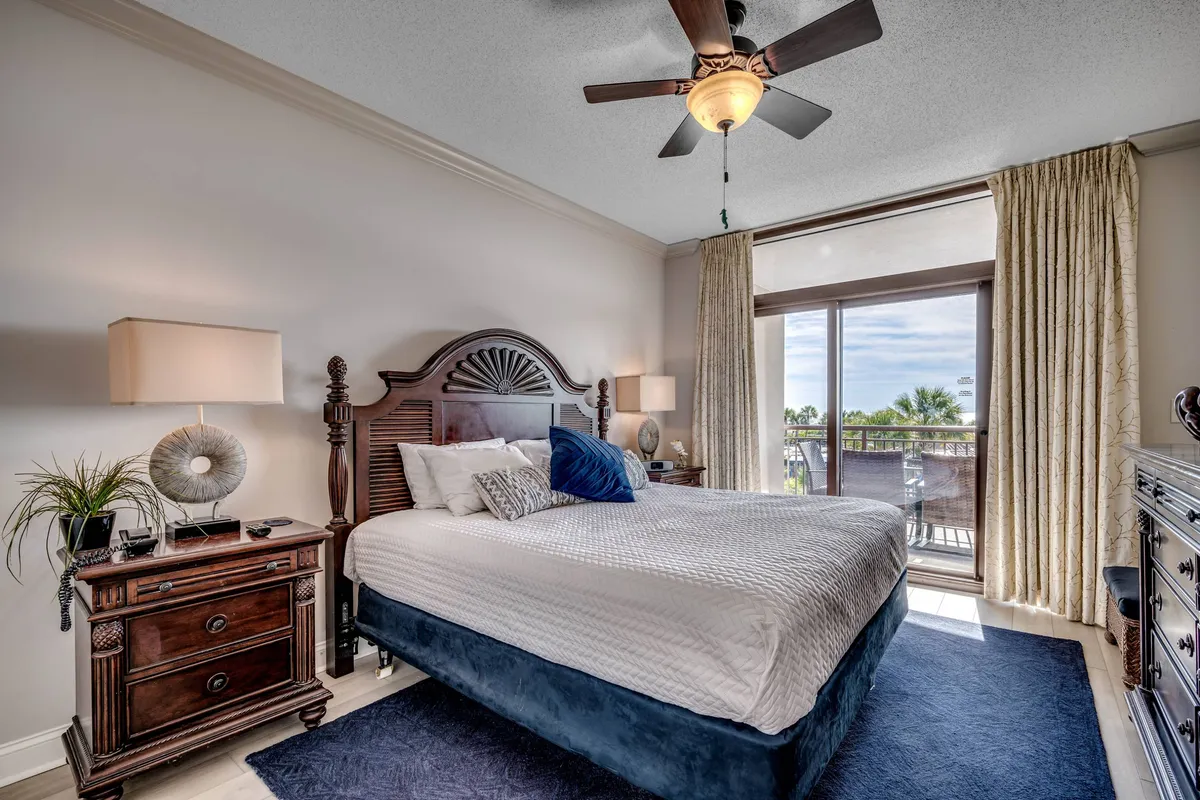 This serene bedroom features a king-sized bed dressed in crisp white linens with decorative throw pillows. Large windows allow natural light to fill the space, while a wooden nightstand and ceiling fan add to the comfortable, modern atmosphere. The overall look is calm and inviting, in line with a restful retreat.