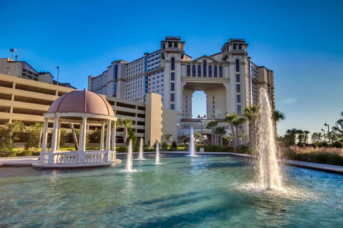 An elegant outdoor scene featuring a fancy gazebo overlooking water fountains, with the large resort building visible in the background. The area is bathed in golden sunset light, inviting guests to relax by the water or enjoy a peaceful stroll.