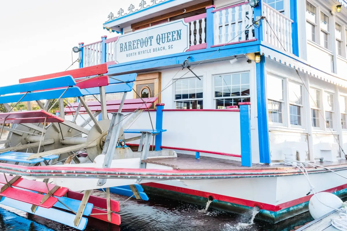 A vibrant paddlewheel boat docked by the waterfront with colorful exterior details and signage, inviting groups for boat tours or casual waterfront dining. The scene hints at live music, social events, and unique group dining experiences, with a relaxed yet lively mood.