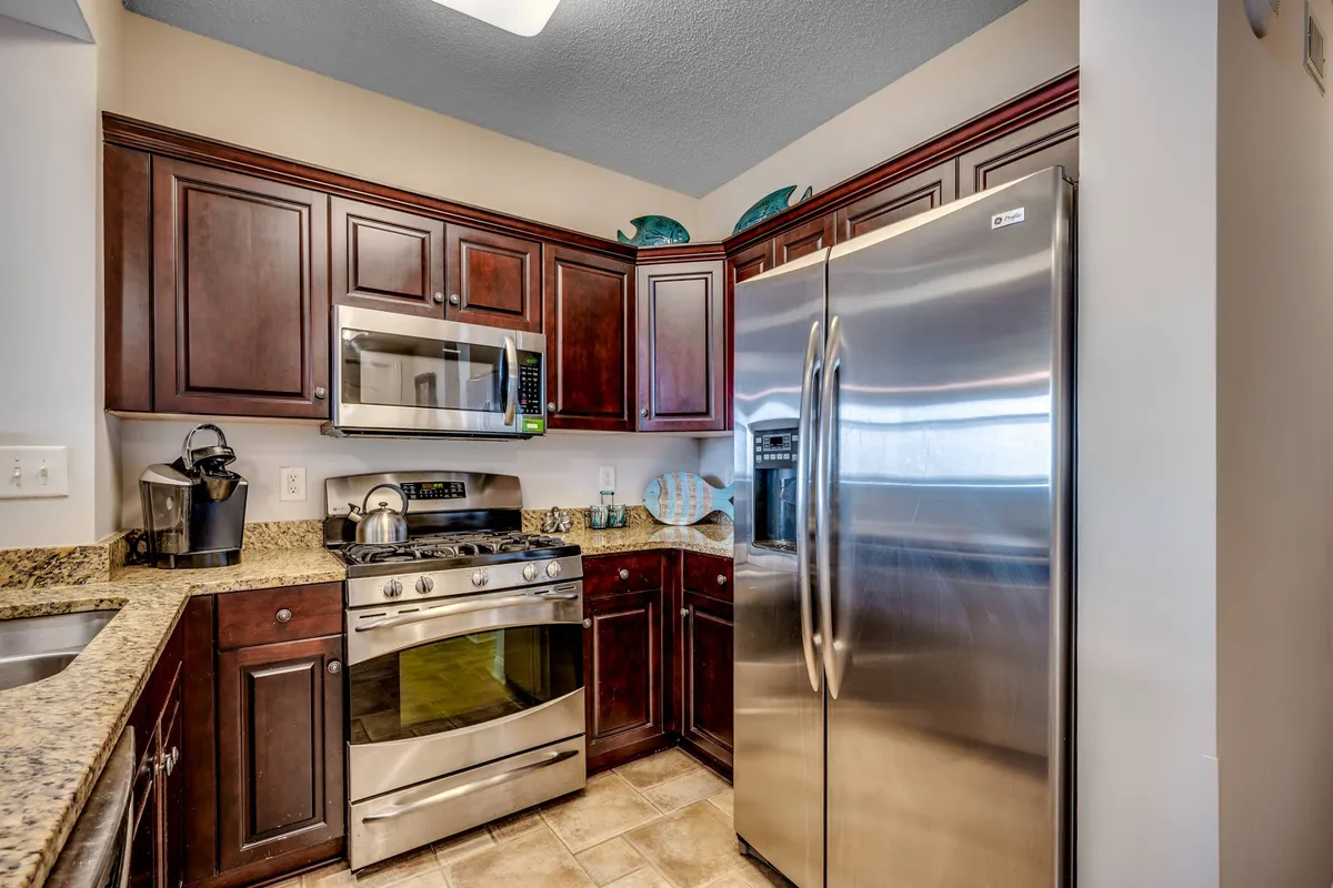 A welcoming chef’s kitchen with dark wooden cabinets, sleek granite countertops, and a full suite of modern stainless steel appliances, including a refrigerator, oven, and microwave. Small appliances like a coffee maker and blender are visible on the counters, while the spacious layout invites easy movement and conversation.