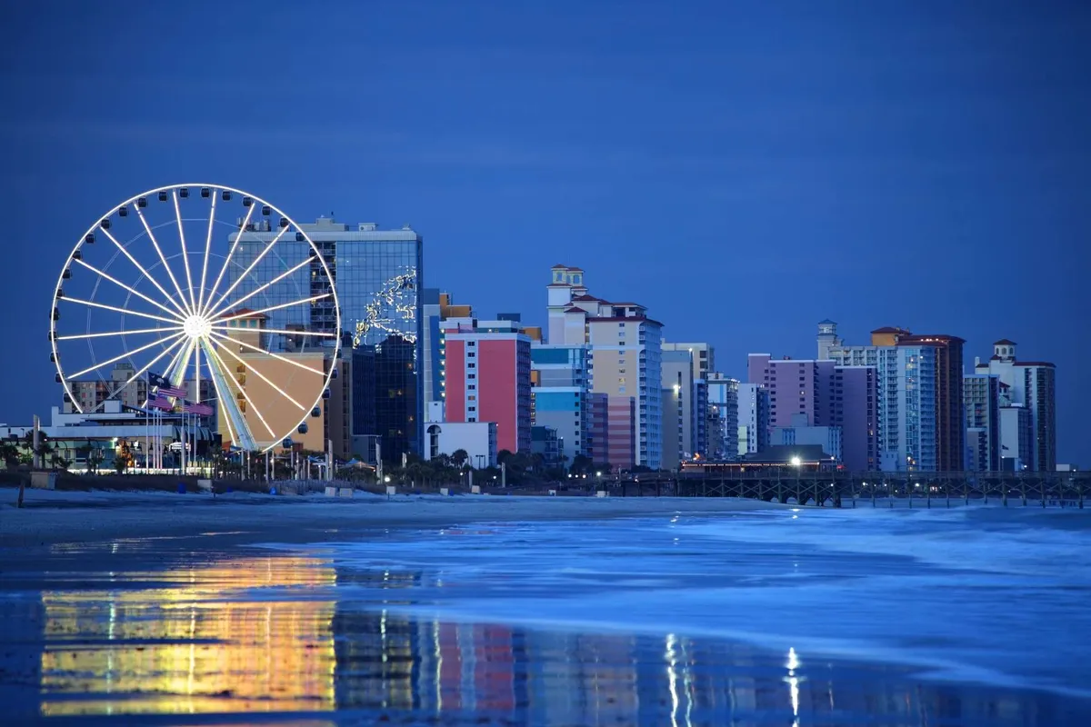 A vibrant coastal scene featuring a towering Ferris wheel, colorful buildings, and reflections of lights on the ocean at sunset. The setting is lively and playful, inviting families to explore amusement rides and oceanfront dining as day turns to night.