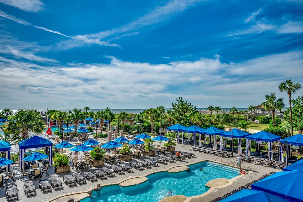 A lively and expansive outdoor resort pool area with a large swimming pool, surrounded by palm trees, beach umbrellas, and plenty of lounge chairs. The scene evokes a sense of fun and relaxation, with families and groups enjoying the sun, swimming, and socializing. The view hints at the proximity to the beach, with glimpses of the ocean on the horizon.
