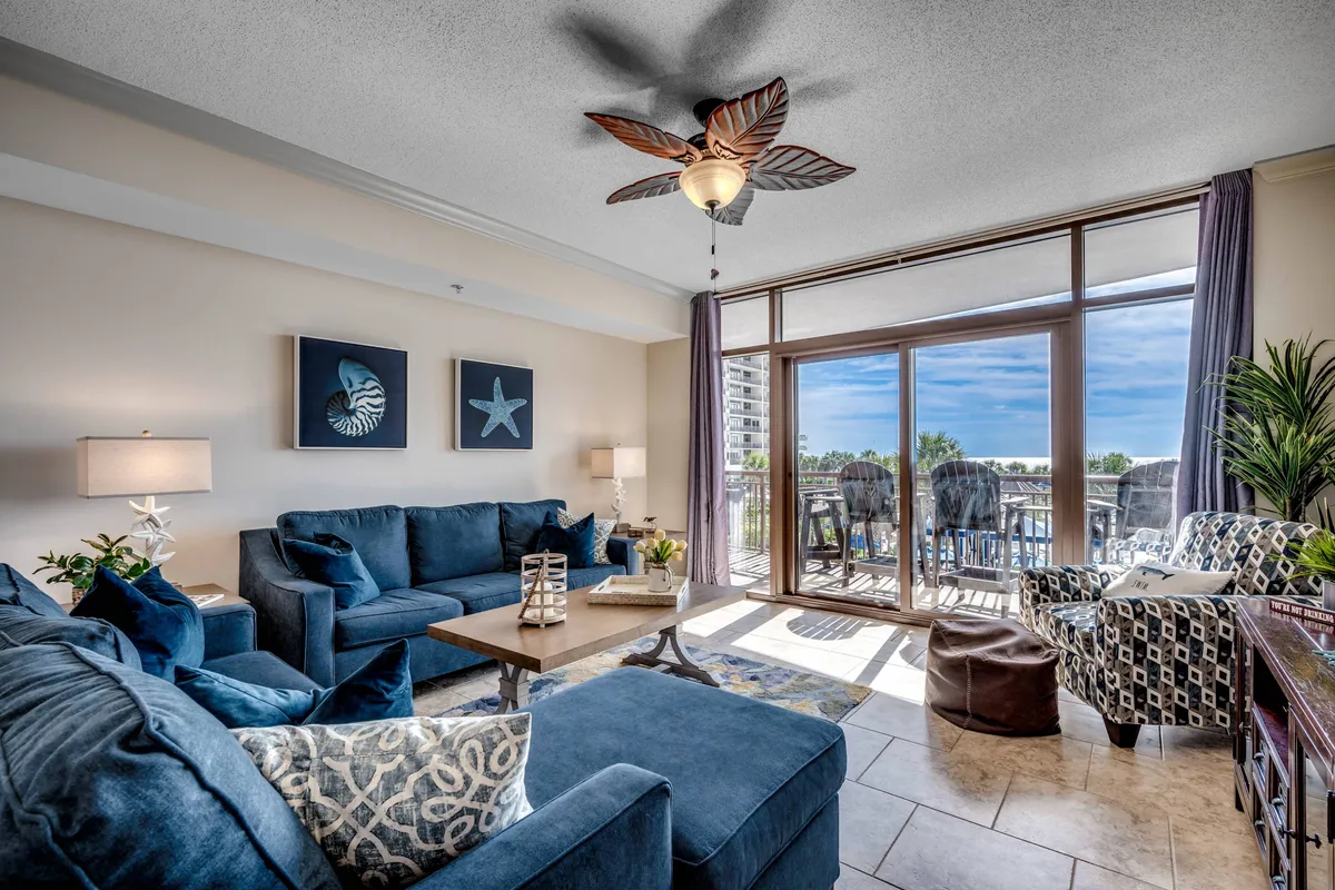 A cozy living room with a plush sofa and coffee table sits adjacent to a set of sliding doors, which open to reveal a private balcony with panoramic ocean views. The room is bright and airy, with sunlight streaming in and a ceiling fan overhead, encouraging relaxation and socializing.