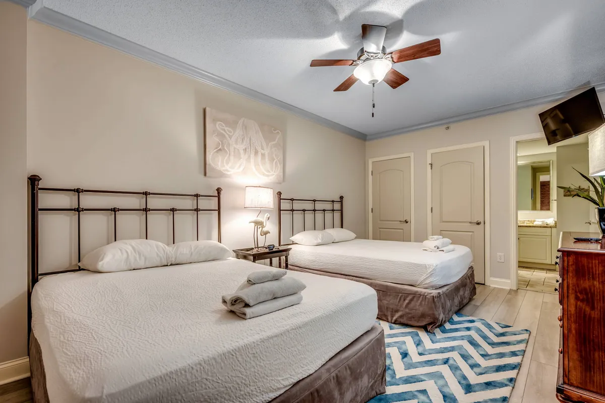 The guest bedroom is shown with two queen-sized beds, both featuring white bedding and decorative wall art above. A wooden dresser and a ceiling fan complete the space, which exudes a relaxed and welcoming mood perfect for friends or families needing extra sleeping options.
