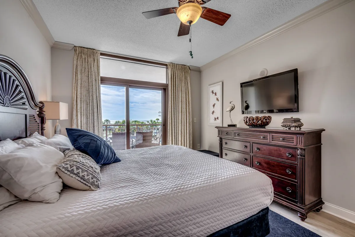 A serene bedroom interior with a bed dressed in decorative pillows, a wooden dresser, and a large window offering an impressive view—likely of the coast or resort grounds. The space exudes tranquility and comfort, suggesting the pleasure of waking up to beautiful scenery.