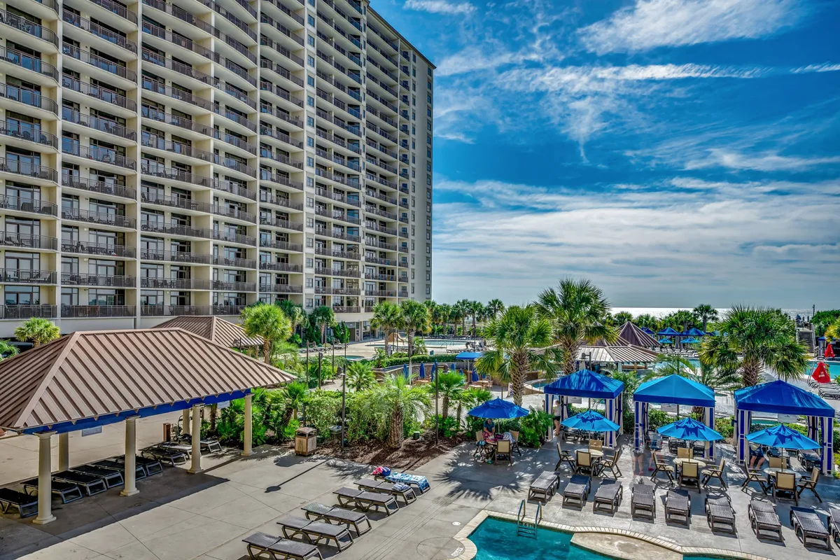 An expansive pool area with blue umbrellas, lounge chairs, and swaying palm trees is set beside a tall resort building. Children and adults are swimming and playing, creating a joyful, vacation-like scene perfect for families.
